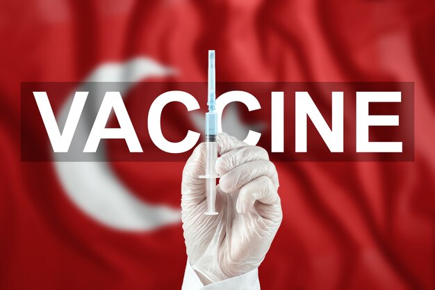 Strzykawka ze szczepionką w dłoni lekarza na tle flagi tureckiej. Koronawirus COVID-19, ochrona immunologiczna, leczenie przeciwko zakażeniu nCoV 2019, szczepienia.