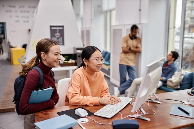 Studenci w bibliotece uczelni skupiają się na dwóch uśmiechniętych młodych dziewczynach korzystających z komputera