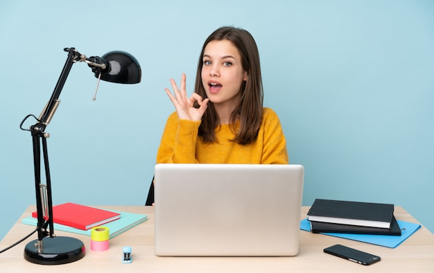 Studencka dziewczyna studiuje w jej domu na błękit ścianie zaskakującej i pokazuje ok znaka