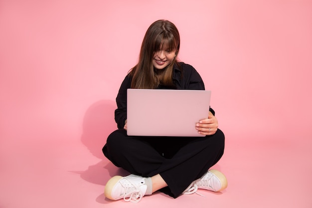 Student Brown Haired zaskoczony uroczy Dziewczyna w dorywczo ubrania Holding Laptop Siedzi Na Różowym Tle. Koncepcja edukacji online. Strzał studio. Patrząc na laptopa.