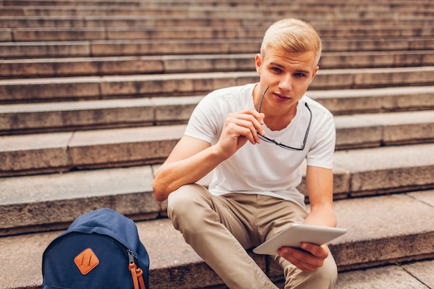 Student collegu z plecakiem używać pastylki obsiadanie na schodkach i mienia szkłach. Facet studiuje na zewnątrz