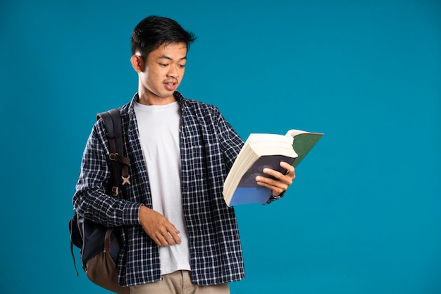 Student mężczyzna ubrany w koszulę zdumiony, trzymając książkę na białym tle na niebieskim tle