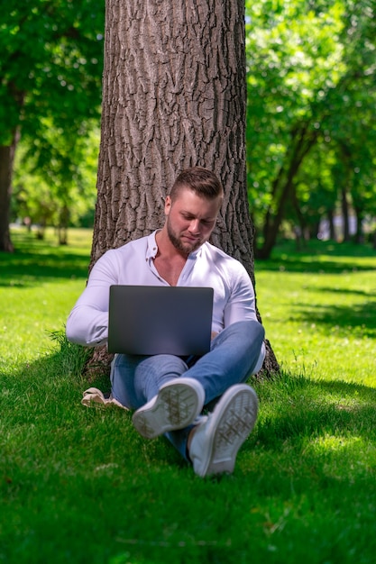 Student ogląda wideo na laptopie siedząc na trawie obok drzewa w wiejskim parku w słoneczny dzień