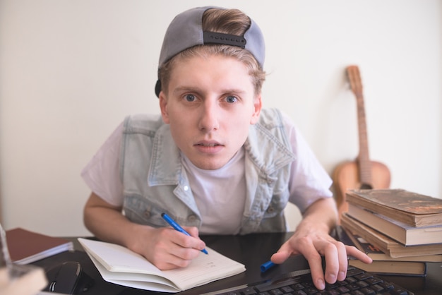 Student siedzi przy komputerze w pobliżu książek, patrzy na monitor i pisze w zeszycie.