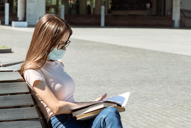 Student w masce z książkami i plecakiem na ławce w parku w letniej ochronie przed covidowymi nastolatkami i dziećmi
