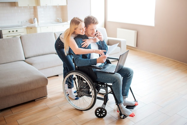 Student z niepełnosprawnością siedzący na wózku inwalidzkim. Wesoła kobieta stoi za nim i obejmuje go. Patrząc na laptopa. Młody człowiek ze specjalnymi potrzebami. Para razem
