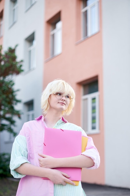 Zdjęcie studentka spaceruje po mieście z folderami na notatnik i stoi przed kampusem uniwersyteckim