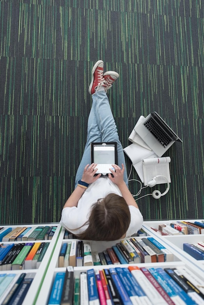 Zdjęcie studentka studiuje w szkolnej bibliotece za pomocą tabletu i szuka informacji w internecie słuchanie muzyki i lekcji na białych słuchawkach