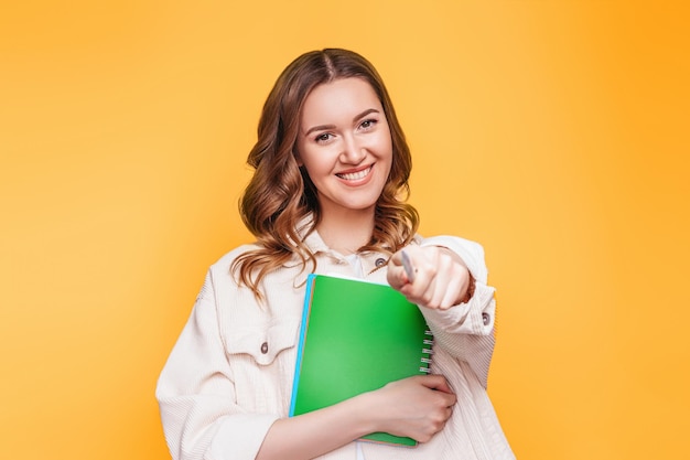 Studentka Uśmiecha Się I Wskazuje Palcem Prosto W Kamerę Przed Sobą Odizolowaną Na Pomarańczowym Tle Miejsca Kopiowania