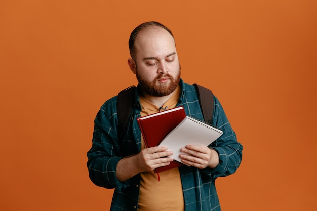 Studentka w codziennych ubraniach z plecakiem trzymająca zeszyty, która wygląda pewnie z poważnym wyrazem twarzy, stojąc na pomarańczowym tle