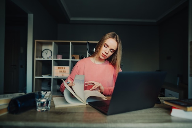 Studentka w różowym swetrze studiuje zdalnie w domu, patrzy w notatniku z poważną miną