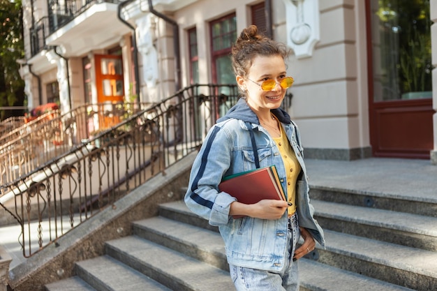 Studentka w średnim wieku, atrakcyjna kobieta z kręconymi włosami i żółtymi okularami przeciwsłonecznymi w stylowych dżinsowych ubraniach, trzymająca książki w mieście w jasny słoneczny dzień