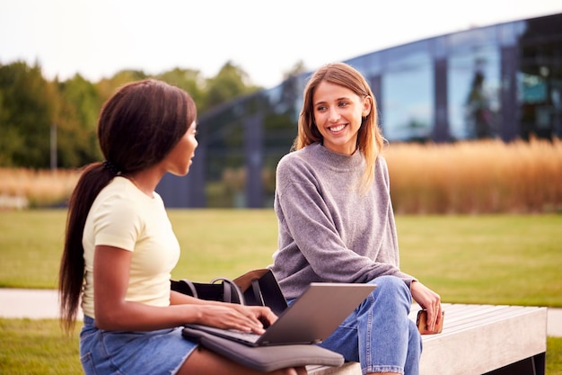 Studentki uniwersytetu lub college'u siedzą na zewnątrz na kampusie rozmawiają i pracują na laptopie