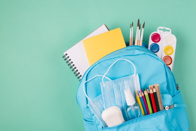 Studia podczas koncepcji kwarantanny. Widok z góry na zdjęcie plecaka wypełnionego kolorową papeterią, dwiema niebieskimi maskami, mydłem i środkiem do dezynfekcji rąk, na białym tle na turkusowym tle