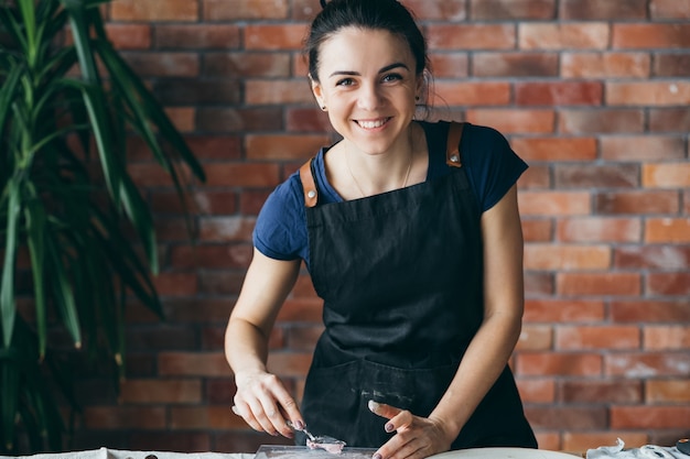 Studio artystyczne. Rękodzieło z gliny w procesie. Uśmiechnięta młoda kobieta z narzędziami do modelowania w miejscu pracy, patrząc na kamery.