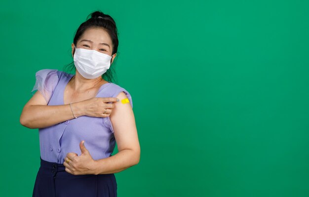 Studio bliska strzał azjatyckich kobiet pacjenta nosić maskę patrzeć na aparat pokazując kciuk z żółtym bandażem gipsu po otrzymaniu szczepienia koronawirusa covid 19 od lekarza na zielonym tle.