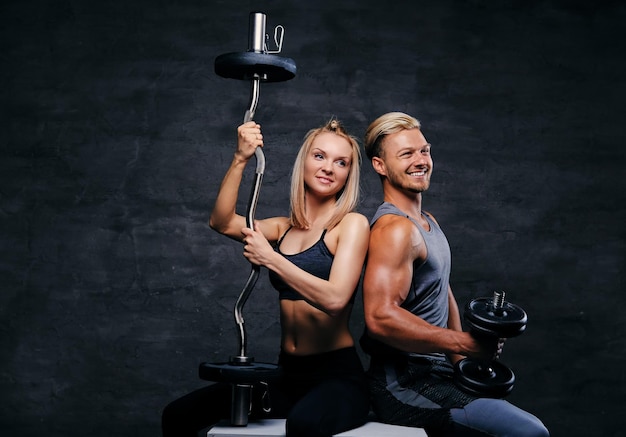 Studio portret atrakcyjna para fitness posiada sztangę i hantle, siedzieć na drewnianym pudełku na ciemnoszarym tle.