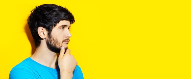 Studio portret młodego rozważnego faceta dotykając brody, patrząc na puste tło żółtego koloru z miejsca na kopię.
