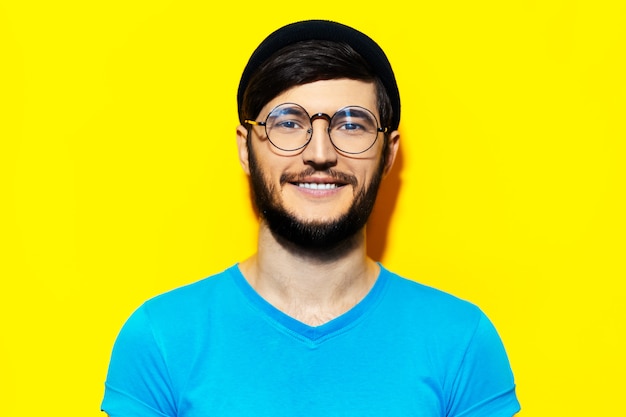 Studio portret młodego uśmiechniętego faceta hipster na sobie kapelusz beanie, okrągłe okulary i niebieską koszulę na tle żółtego koloru.