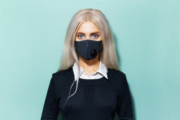 Studio portret młodej blondynki o niebieskich oczach w szkolnej formie, ubrana w czarną maskę oddechową przeciwko koronawirusowi na tle koloru cyjan, aqua menthe.