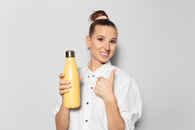 Studio Portret Młodej Uśmiechniętej Dziewczyny Trzymającej Termofor W Kolorze żółtym Na Białym Tle Pokazuje Gest Kciuka W Górę