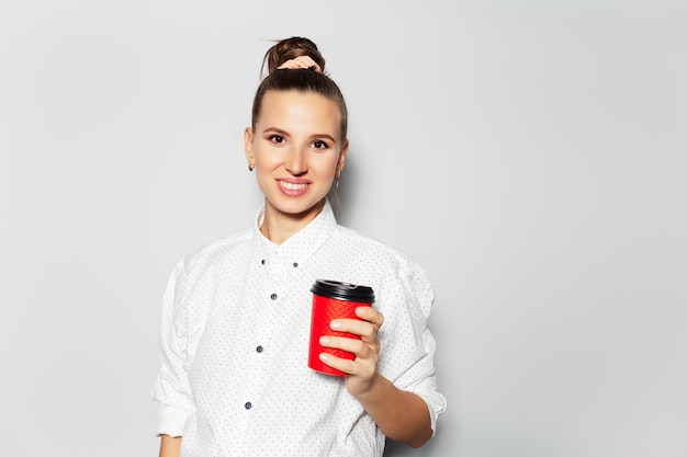 Studio portret młodej uśmiechniętej dziewczyny trzymającej w ręku papierowy kubek na kawę na wynos na białym tle