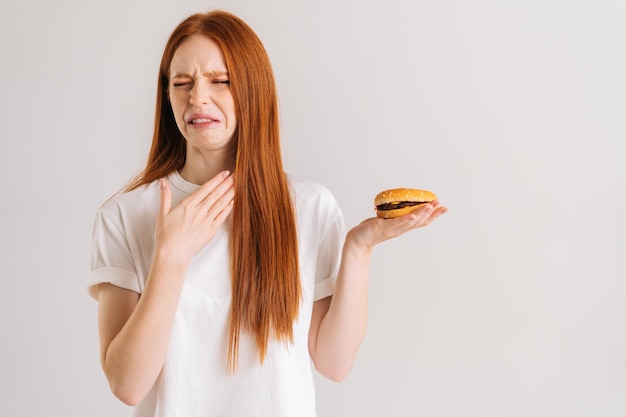 Studio portret niezadowolonej ładnej młodej kobiety z obrzydzeniem wskazującym na zamknięte oczy burgera