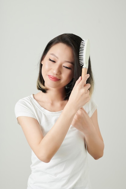 Studio portret pozytywnej młodej azjatyckiej kobiety cieszącej się szczotkowaniem włosów