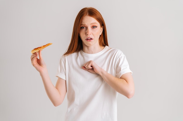 Studio Portret Przestraszonej Młodej Kobiety Cierpiącej Na Duszenie I Kaszel Po Zjedzeniu Pizzy, Bije Się W Klatkę Piersiową, Stojąc Na Białym Tle Na Białym Tle, Patrząc Na Kamerę, Trzymając Kawałek Pizzy.