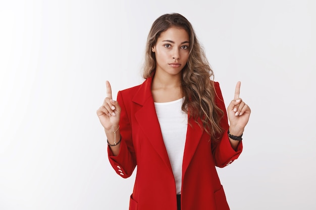 Studio strzał zmartwiony niezdecydowany młody absolwent żeński pracownik biurowy ubrany w czerwoną kurtkę konsultujący współpracownik wskazujący palce wskazujące w górę patrzący pytany niepewny poważna twarz, stojąca biała ściana