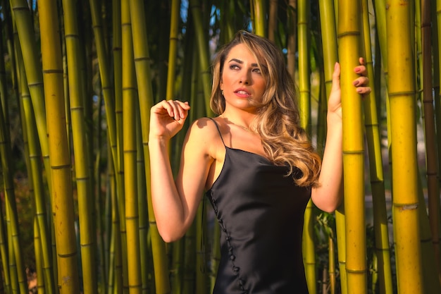 Styl życia blondynka kaukaska dziewczyna w czarnej sukience na ramiączkach w pięknym bambusie w parku na świeżym powietrzu dziewczyna siedząca trzymając się słupa i patrząc w lewo