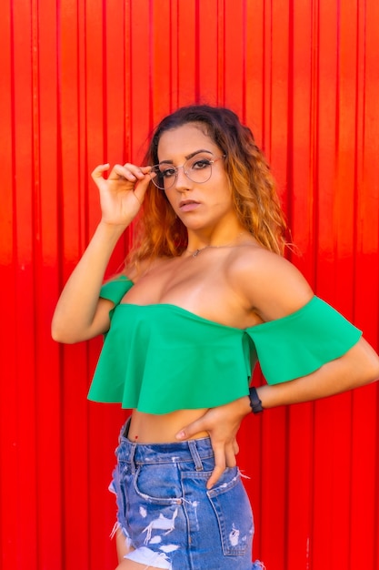 Styl życia Kaukaska blondynka w zielonych koszulkach dżinsowych szortach i przezroczystych okularach na czerwonej ścianie Nastoletnia studentka w letniej pozie