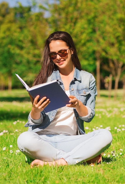 styl życia, letnie wakacje, edukacja, literatura i koncepcja ludzi - uśmiechnięta młoda dziewczyna czytająca książkę i siedząca na trawie w parku