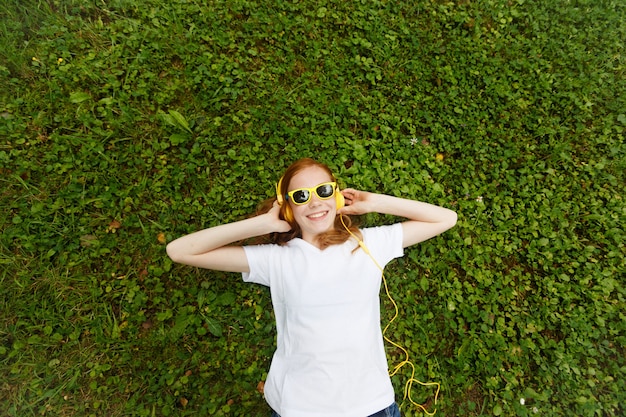 Styl życia, wakacje, technologia, muzyka i koncepcja ludzi - uśmiechnięta młoda dziewczyna z rudymi włosami i słuchawkami, leżąca z zamkniętymi oczami na trawie.