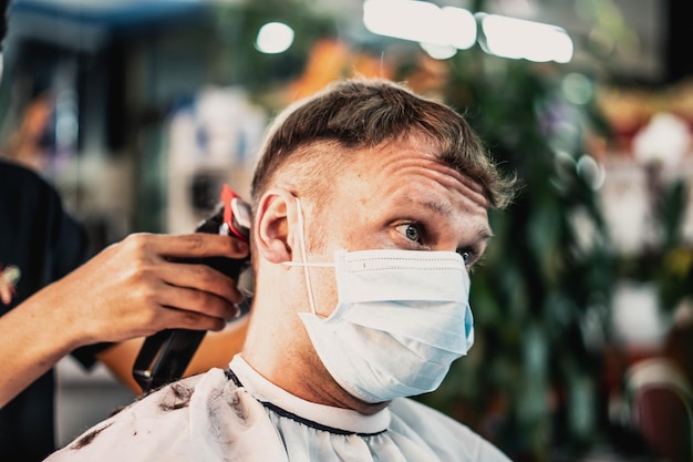 Styl życia zdjęcie fryzjer Młody mężczyzna klient nosi maskę zaskoczony niezadowolony wygląd pomarszczone czoło Fryzjer mężczyzna praca ręczna grzebień narzędzie elektryczna maszynka do strzyżenia Krótkie kosmyki włosów