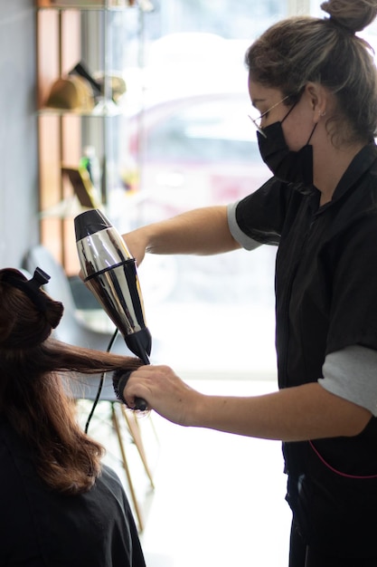 Zdjęcie stylistka przeczesuje włosy swoich klientów suszarką i okrągłym grzebieniem w salonie z dużą ilością naturalnego światła