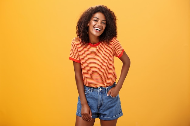 stylowa African American kobieta w szortach i koszulce śmiejąca się radośnie nad pomarańczową ścianą