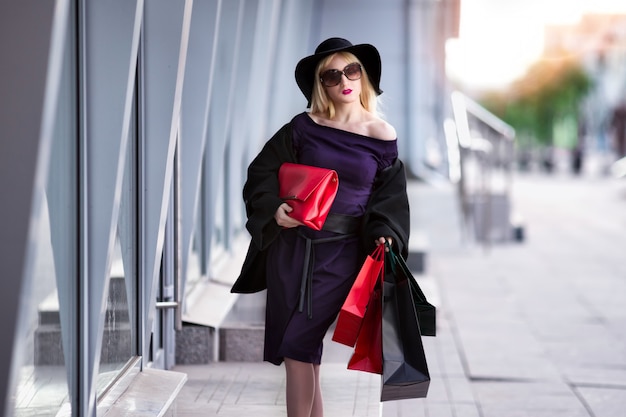 Stylowa blondynka w czarnym płaszczu, okularach przeciwsłonecznych i kapeluszu z torbami na zakupy spaceruje ulicą, świąteczny koncert