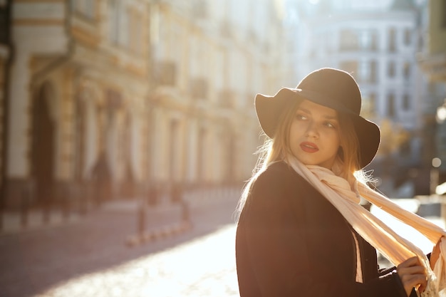 Stylowa blondynka w kapeluszu, szaliku i płaszczu, idąc starą ulicą w promieniach słońca. Miejsce na tekst