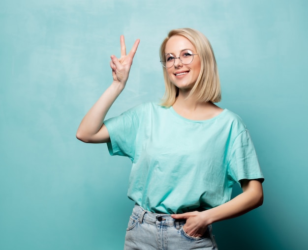 Stylowa blondynki kobieta w szkłach pokazuje OK gest na błękit ścianie