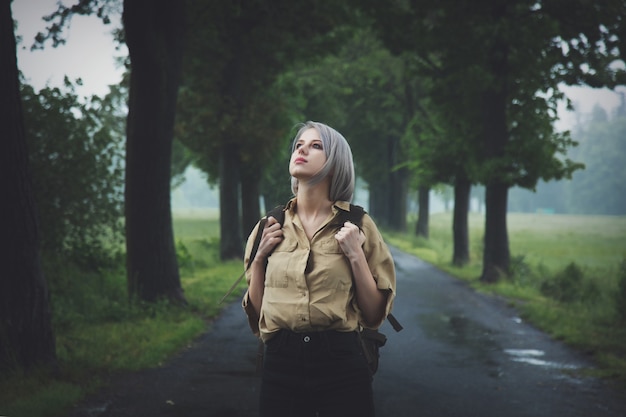 Stylowa blondynki kobieta z plecakiem przy wsi drogą w deszczowym dniu