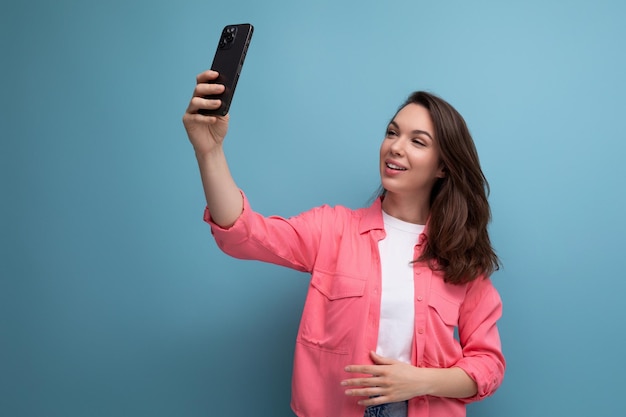 Stylowa brunetka kobieta w modnej koszuli robi sobie selfie za pomocą telefonu na tle studia