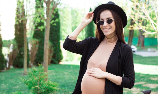 Stylowa ciąża w kapeluszu i okularach przeciwsłonecznych siedząca na ławce w parku miejskim i relaksująca