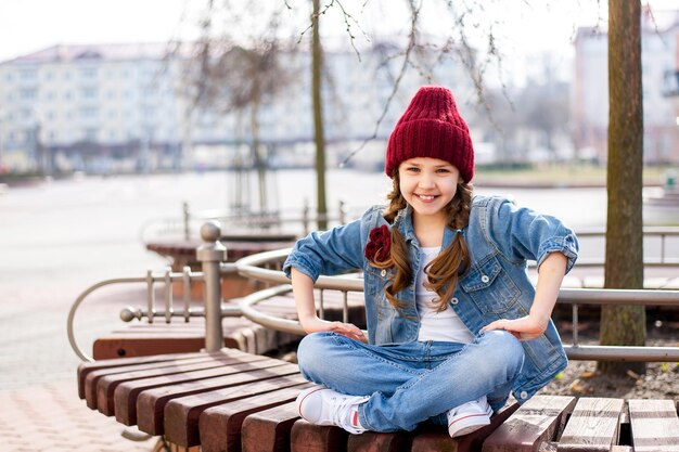 Zdjęcie stylowa dziewczyna siedzi na ławce na ulicy i uśmiecha się do fotografa
