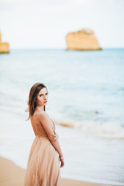stylowa dziewczyna stojąca z rozwianymi włosami od wiatru o zachodzie słońca na plaży i oceanie