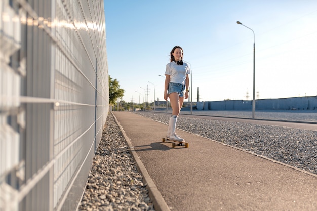 Stylowa dziewczyna w białych pończochach jeździ na longboardzie ulicą i słucha muzyki