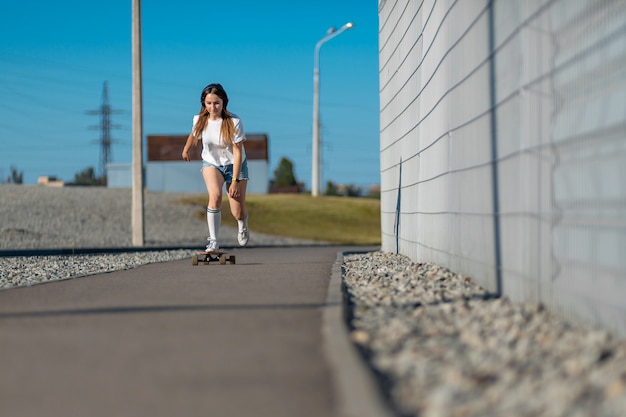 Stylowa dziewczyna w białych pończochach jeździ na longboardzie ulicą i słucha muzyki