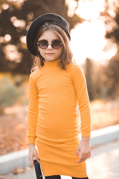 Stylowa dziewczynka 56-letnia nosi żółtą sukienkę i beret z okularami przeciwsłonecznymi w jesiennym parku na zewnątrz Spójrz na kamerę Sezon jesienny Dzieciństwo