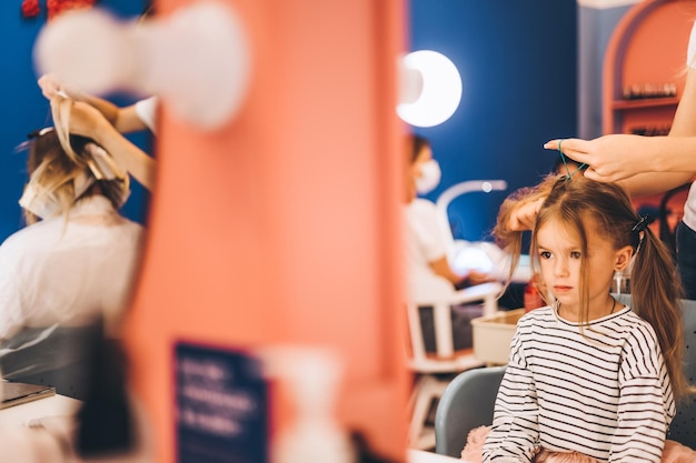 Stylowa dziewczynka w salonie kosmetycznym dla dzieci, gdzie wykonała piękną fryzurę Stylistka robi stylową fryzurę małej dziewczynce