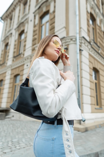 Zdjęcie stylowa hipster ładna dziewczyna w modnych ubraniach ze skórzaną kurtką i torebką nosi żółte okulary i spaceruje po ulicy